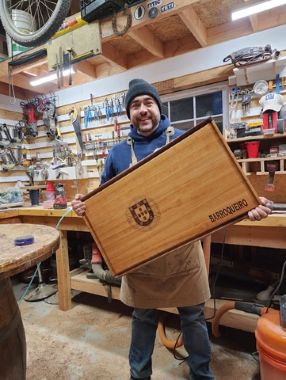 A woodworker at Iron Shed Woodworks holding a finished wood box in the workshop, surrounded by tools and workbenches