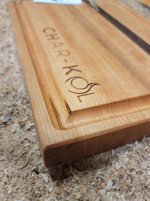 Cherry board with a deep juice groove and a Char-Kol logo engraved into the face, resting in shop sawdust
